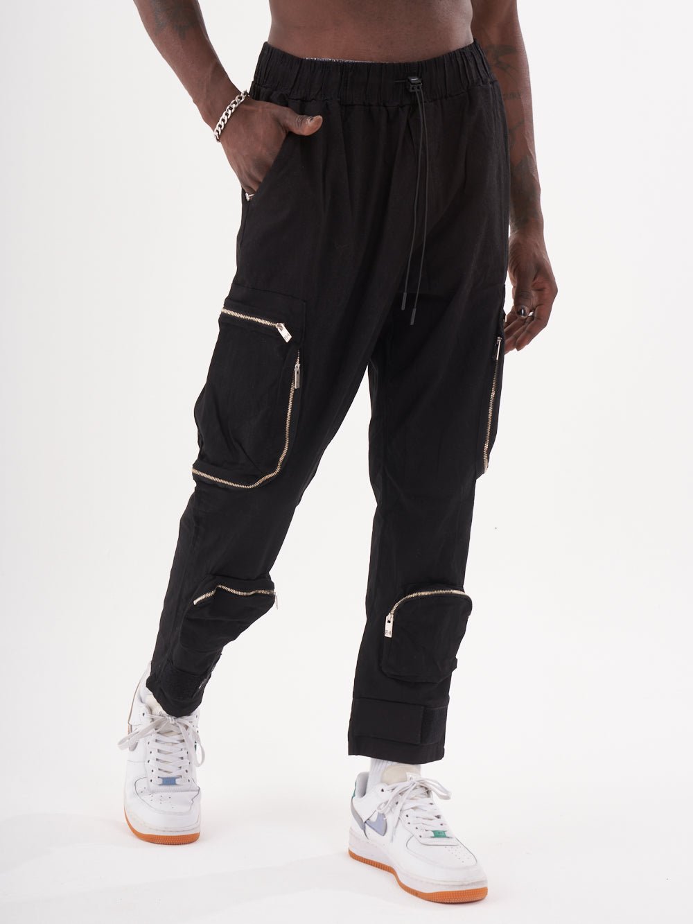 A person is wearing the MULTI-ZIPPER POCKET JOGGER // BLACK with multiple zippered pockets and white sneakers, showcasing the jogger comfort and featuring an elastic drawstring waist for a snug fit.