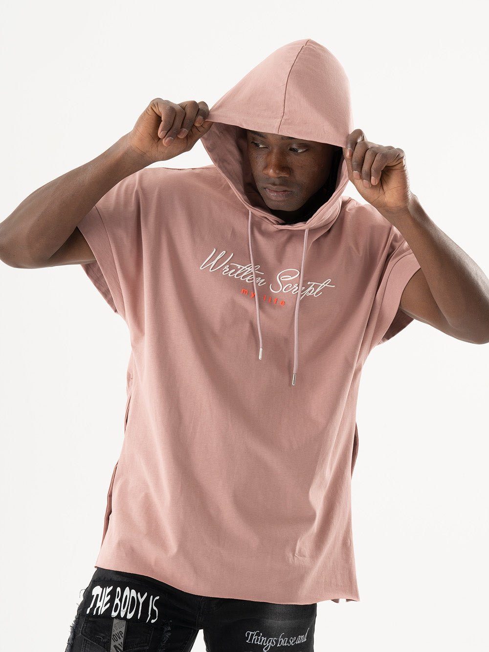 A man wearing a salmon short-sleeve RIGGED HOODIE with the text "Written Script" on the front, pulling the hood over his head. The comfort-fit hoodie matches his black pants adorned with white text patches, creating a casual and stylish look.